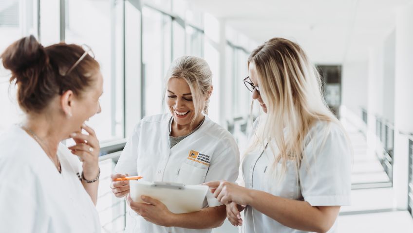 Drei lachende Mitarbeiterinnen von NEURO Südwestfalen in weißen Kasacks in einem Gang des Albertus Magnus Zentrums in Siegen.