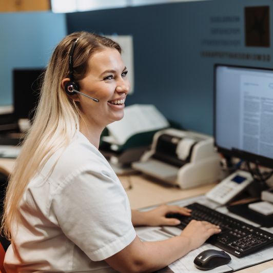 Eine blonde medizinische Fachangestellte sitzt mit Headset an einem Schreibtisch mit Computer, ihre Hände liegen auf der Tastatur. Sie lächelt und arbeitet vermutlich am Empfang.