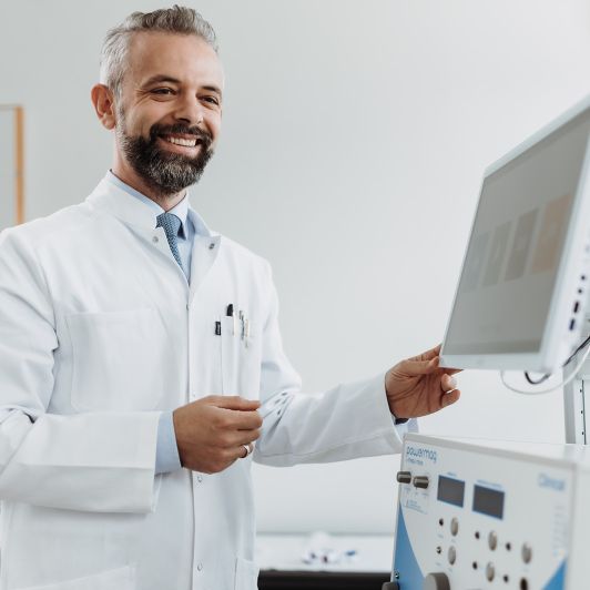 Dr. Gjergji Sinani, Facharzt für Psychiatrie und Psychotherapie, trägt einen weißen Arztkittel und blickt lächelnd auf den Monitor eines Behandlungsgeräts.