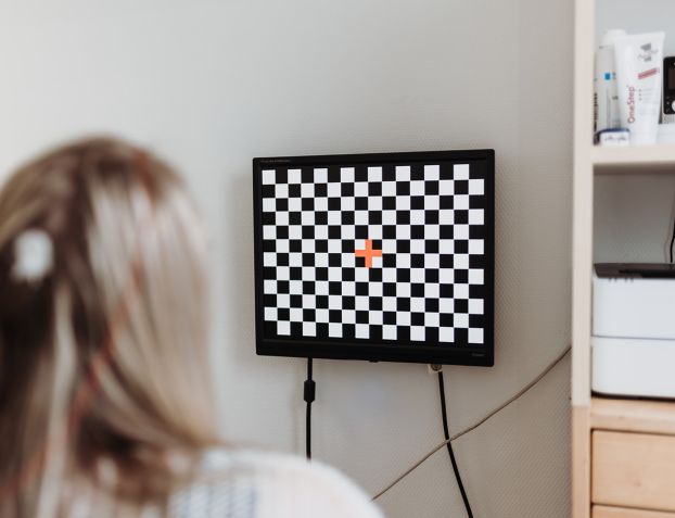 Eine Person mit langen Haaren sitzt vor einem Monitor, der ein schwarz-weißes Schachbrettmuster mit einem roten Kreuz in der Mitte anzeigt. Es handelt sich um einen neurologischen Test in der Praxis.