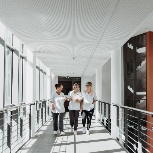 Drei lachende Mitarbeiterinnen von NEURO Südwestfalen in einem Gang des Albertus Magnus Zentrums in Siegen.