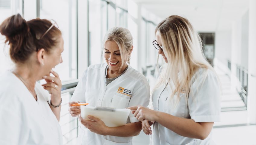 Drei lachende Mitarbeiterinnen von NEURO Südwestfalen in weißen Kitteln, die gemeinsam auf Unterlagen schauen.