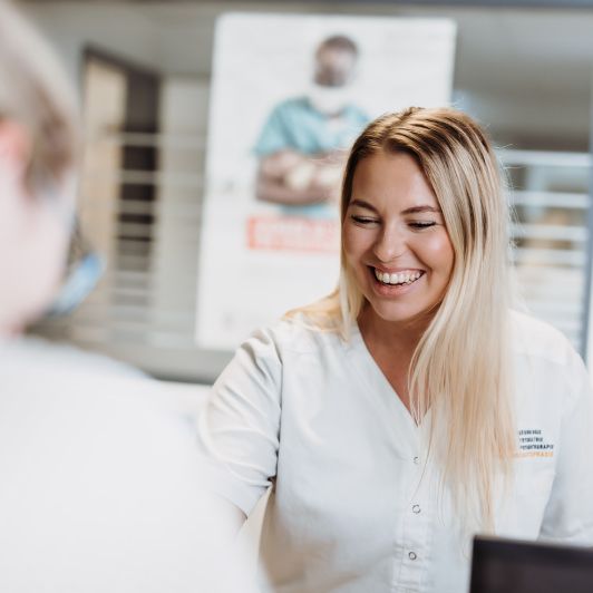 Eine lachende, blonde medizinische Fachangestellte von NEURO Südwestfalen im weißen Kasack, im Gespräch mit einer anderen Person.