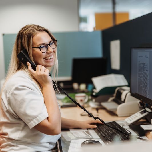 Eine lächelnde, blonde medizinische Fachangestellte mit schwarzer Brille und weißem Kittel sitzt am Computer und hält einen schwarzen Telefonhörer in der Hand.