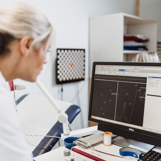 Eine blonde medizinische Fachangestellte im weißen Kittel blickt auf einen Computerbildschirm, auf dem verschiedene Graphen und Messungen einer neurologischen Untersuchung abgebildet sind.