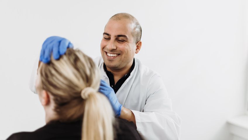 Dr. med. Arya B. Khorsandi lächelt während einer Patienten-Behandlung. Er trägt blaue Handschuhe und untersucht die Patientin am Kopf.