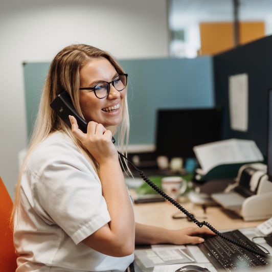 Eine lächelnde, blonde medizinische Fachangestellte mit schwarzer Brille und weißem Kittel sitzt am Computer und hält einen schwarzen Telefonhörer in der Hand.