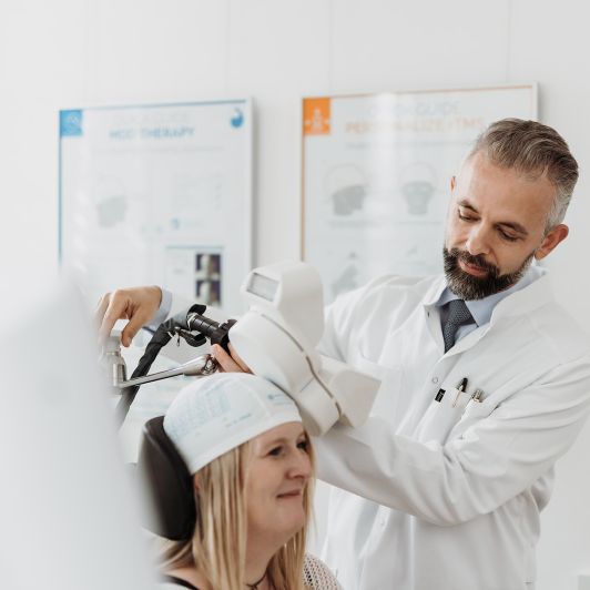 Dr. Gjergji Sinani, Facharzt für Psychiatrie und Psychotherapie, führt eine rTMS-Behandlung (repetitive transkranielle Magnetstimulation) bei einer lächelnden Patientin durch, die eine markierte Kappe trägt.