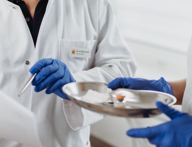 Zwei Mediziner:innen in weißen Kitteln und blauen Einweghandschuhen bereiten ein medizinisches Verfahren vor. Eine Person hält einen Kugelschreiber, während die andere ein silbernes Tablett mit Instrumenten und Materialien trägt.