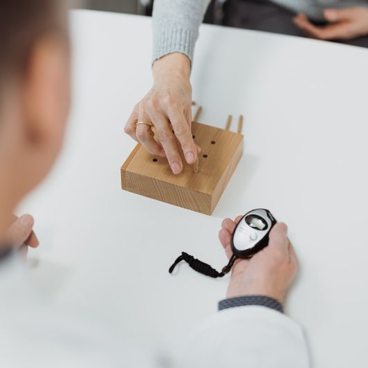 Ein Arzt mit Stoppuhr testet die Reaktionsgeschwindigkeit eines Patienten, der kleine Holzstifte in einen Holzblock mit kleinen Öffnungen einsetzt.
