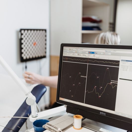 Auf dem Bild sieht man einen Computerbildschirm. Darauf sind verschiedene Graphen und Messungen einer neurologischen Untersuchung zu sehen. Im Hintergrund sieht man eine Patientin.