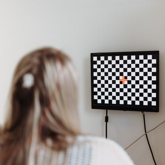 Tests zur Verkehrstauglichkeit Eine Person mit langen Haaren sitzt vor einem Monitor, der ein schwarz-weißes Schachbrettmuster mit einem roten Kreuz in der Mitte anzeigt. Es handelt sich um einen neurologischen Test in der Praxis.