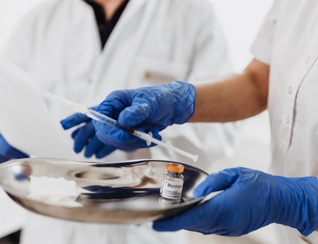 Aufnahme einer medizinischen Fachangestellten, die eine metallene Nierenschale mit einer Ampulle Botulinumtoxin und einer Spritze in der Hand hält.