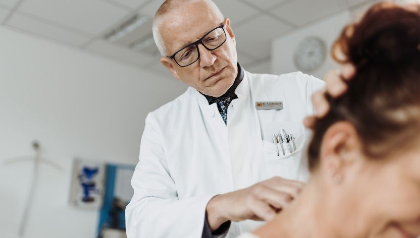 Dr. med. Rainer Lindemuth von NEURO Südwestfalen im weißen Kittel untersucht den Hinterkopf einer braunhaarigen Patientin.