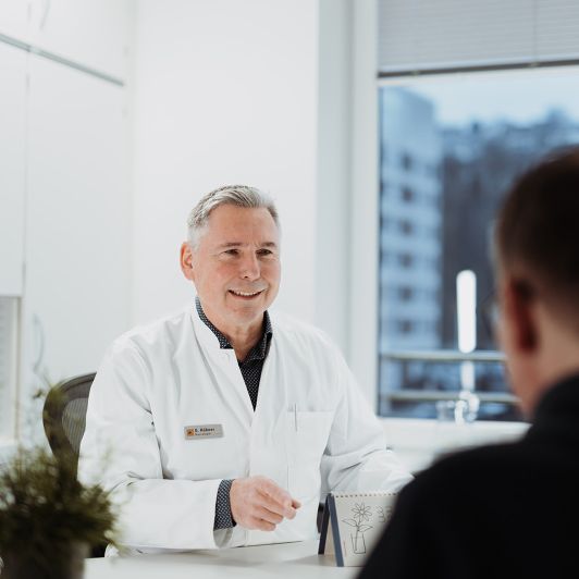 Facharzt für Neurologie und Nervenheilkunde Georg Hübner im weißen Arztkittel, Facharzt für Neurologie und Nervenheilkunde in Siegen und Burbach, im Patientengespräch.