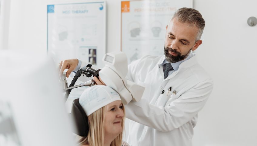 Dr. Gjergji Sinani, Facharzt für Psychiatrie und Psychotherapie, führt eine rTMS-Behandlung (repetitive transkranielle Magnetstimulation) bei einer lächelnden Patientin durch, die eine markierte Kappe trägt.